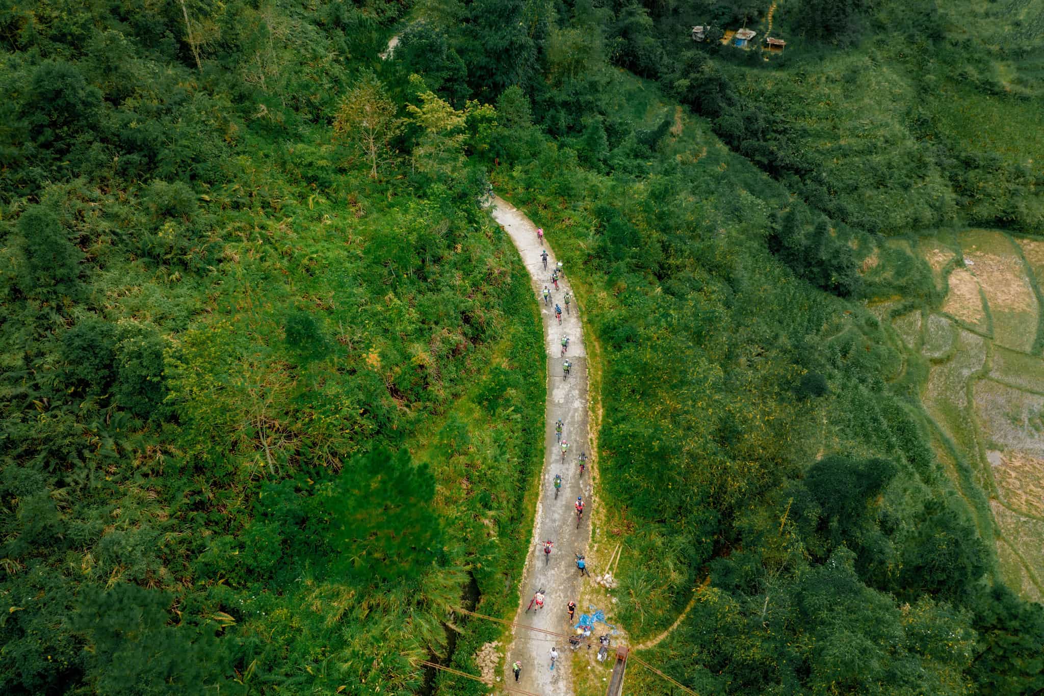 Aerial view of cyclists on a road in Ha Giang, Vietnam, surrounded by forests and fields.