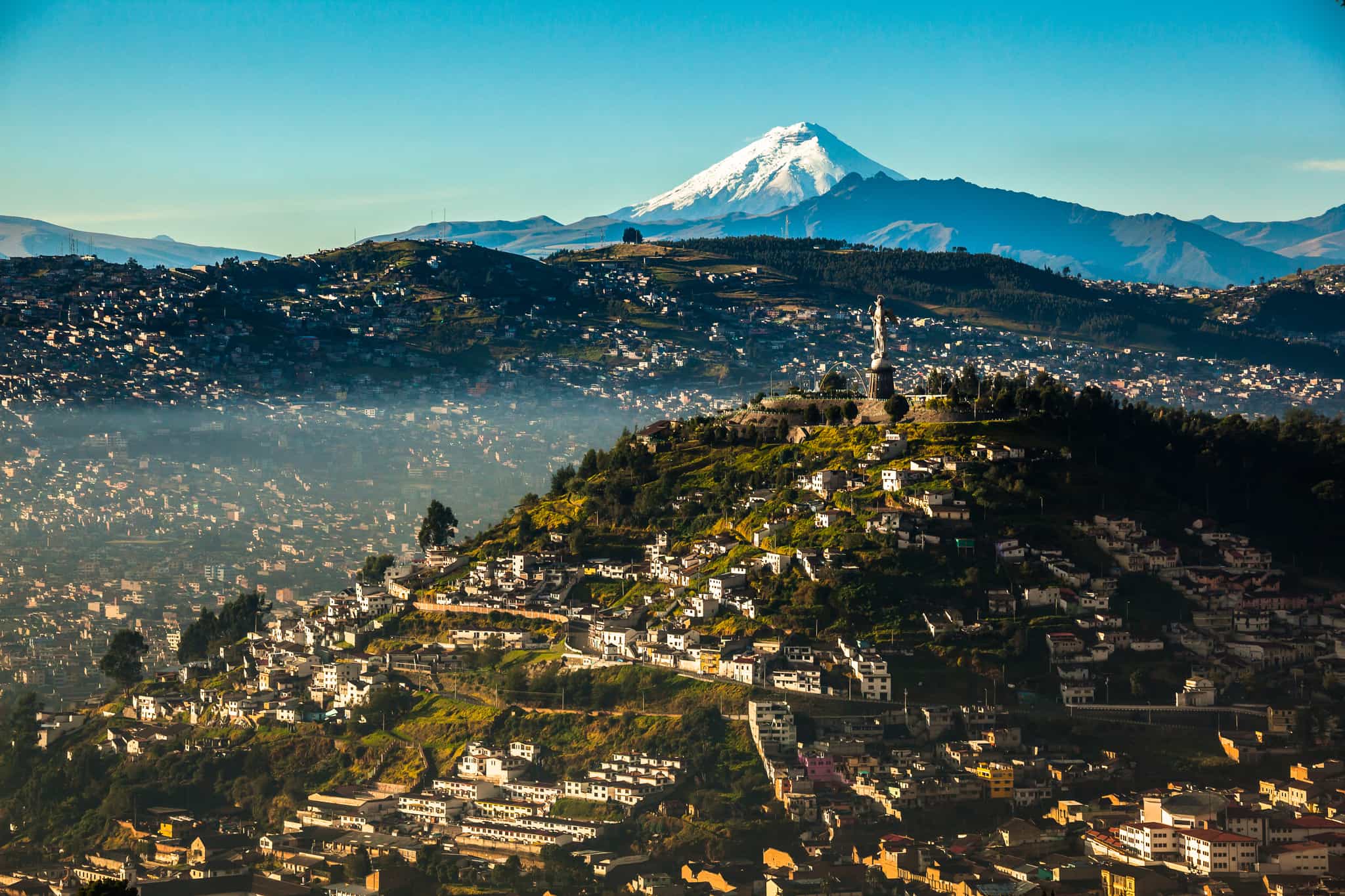A city scape of Quito with the Virgen de El Panecillo statue and Cotopaxi standing proud in the background.